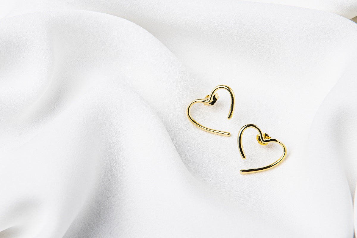 A pair of heart-shaped stud earrings made of sterling silver with gold vermeil, displayed on a white silky surface.