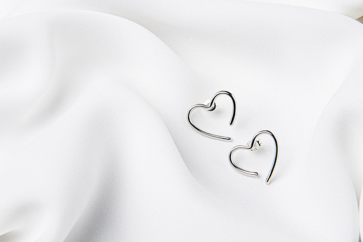 A pair of heart-shaped stud earrings made of sterling silver, displayed on a white silky surface.