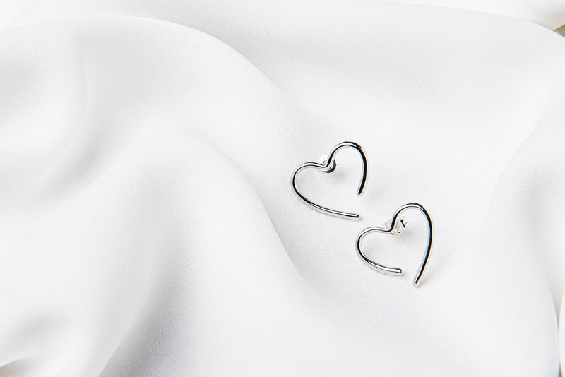 A pair of heart-shaped stud earrings made of sterling silver, displayed on a white silky surface.