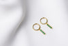 A pair of gold vermeil hoop earrings with a dangle of green cubic zirconias on a white fabric surface.