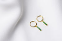 A pair of gold vermeil hoop earrings with a dangle of green cubic zirconias on a white fabric surface.