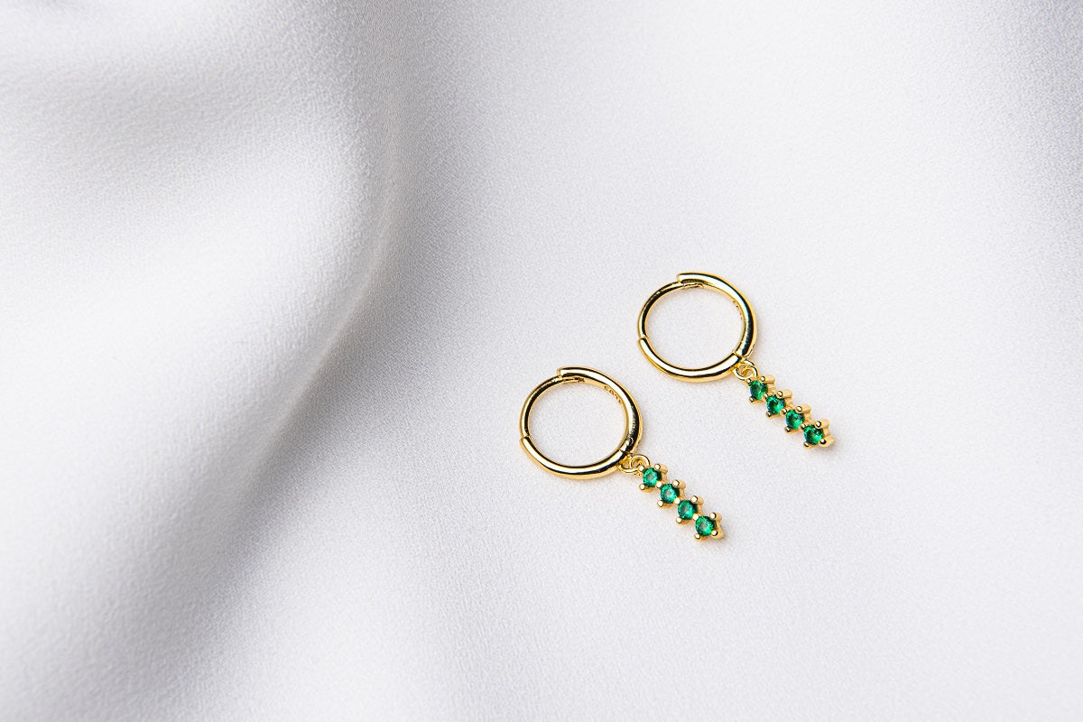 A pair of gold vermeil hoop earrings with a dangle of green cubic zirconias on a white fabric surface.