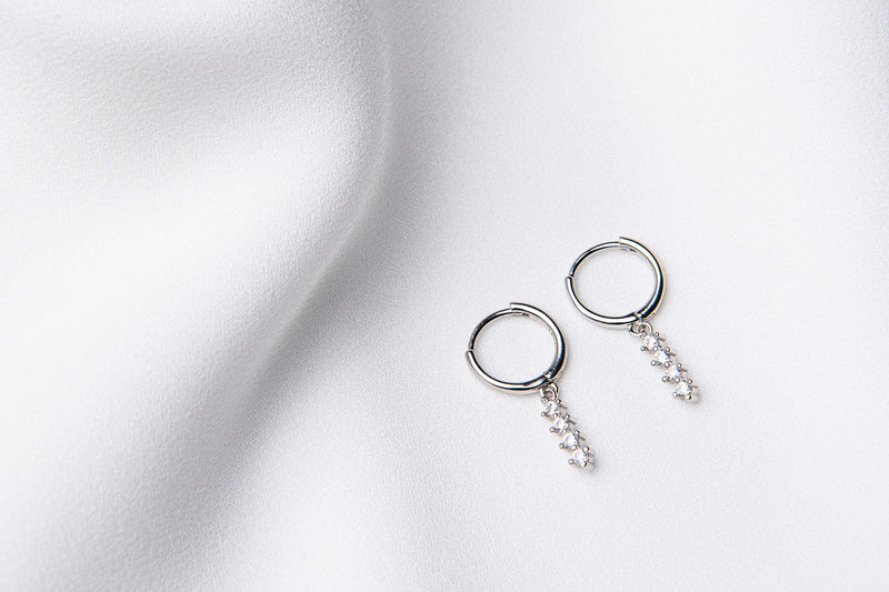 A pair of silver hoop earrings with a dangle of cubic zirconias on a white fabric surface.