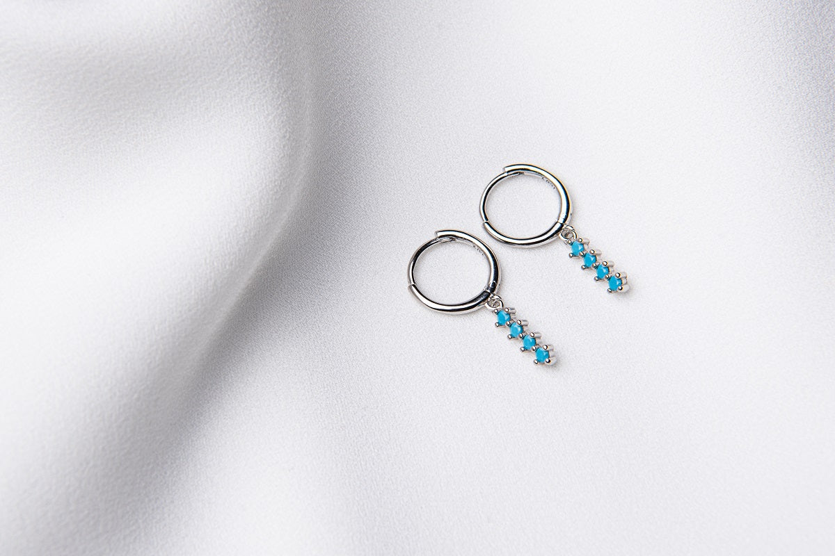 A pair of silver hoop earrings with a dangle of baby blue stones on a white fabric surface.