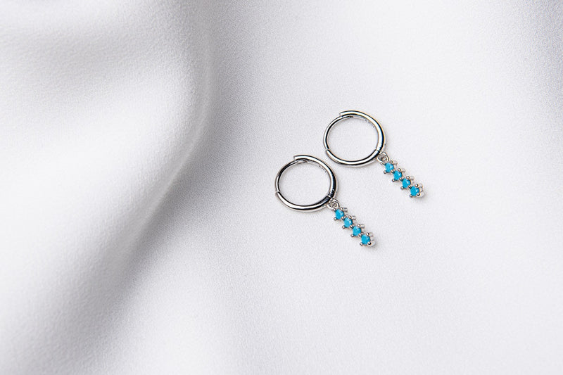 A pair of silver hoop earrings with a dangle of baby blue stones on a white fabric surface.