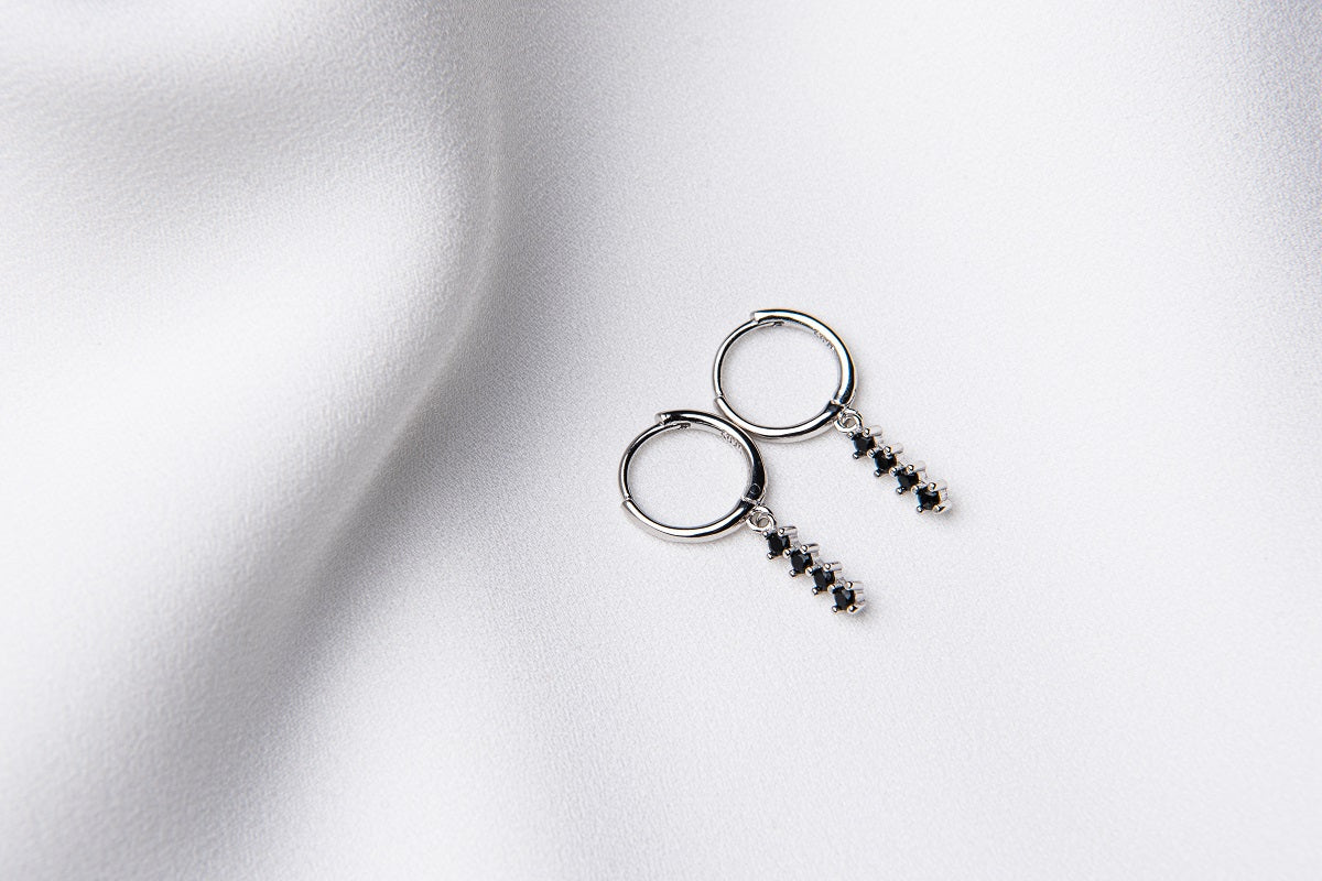 A pair of silver hoop earrings with a dangle of black cubic zirconias on a white fabric surface.