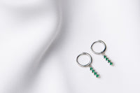 A pair of silver hoop earrings with a dangle of green cubic zirconias on a white fabric surface.
