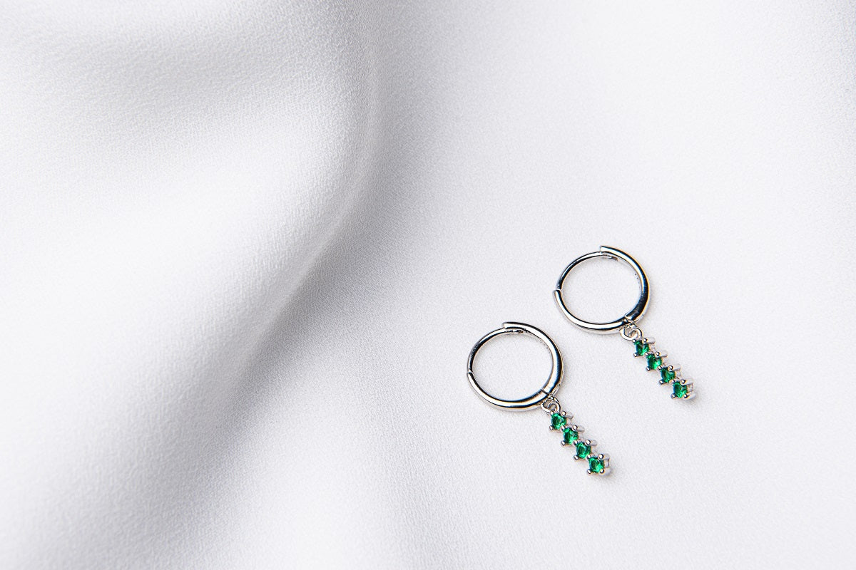 A pair of silver hoop earrings with a dangle of green cubic zirconias on a white fabric surface.