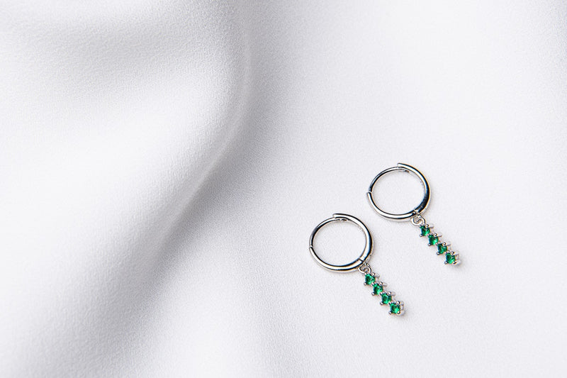 A pair of silver hoop earrings with a dangle of green cubic zirconias on a white fabric surface.