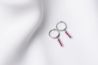 A pair of silver hoop earrings with a dangle of pink cubic zirconias on a white fabric surface.