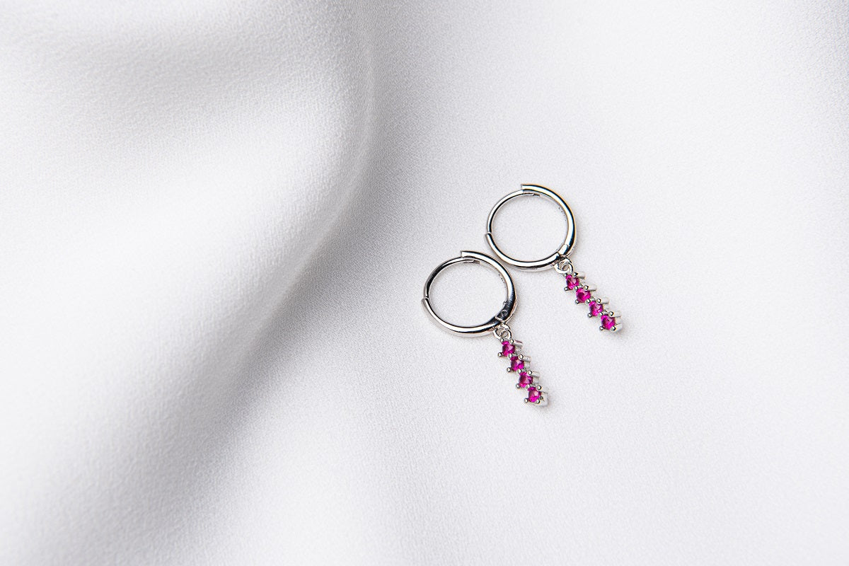 A pair of silver hoop earrings with a dangle of pink cubic zirconias on a white fabric surface.