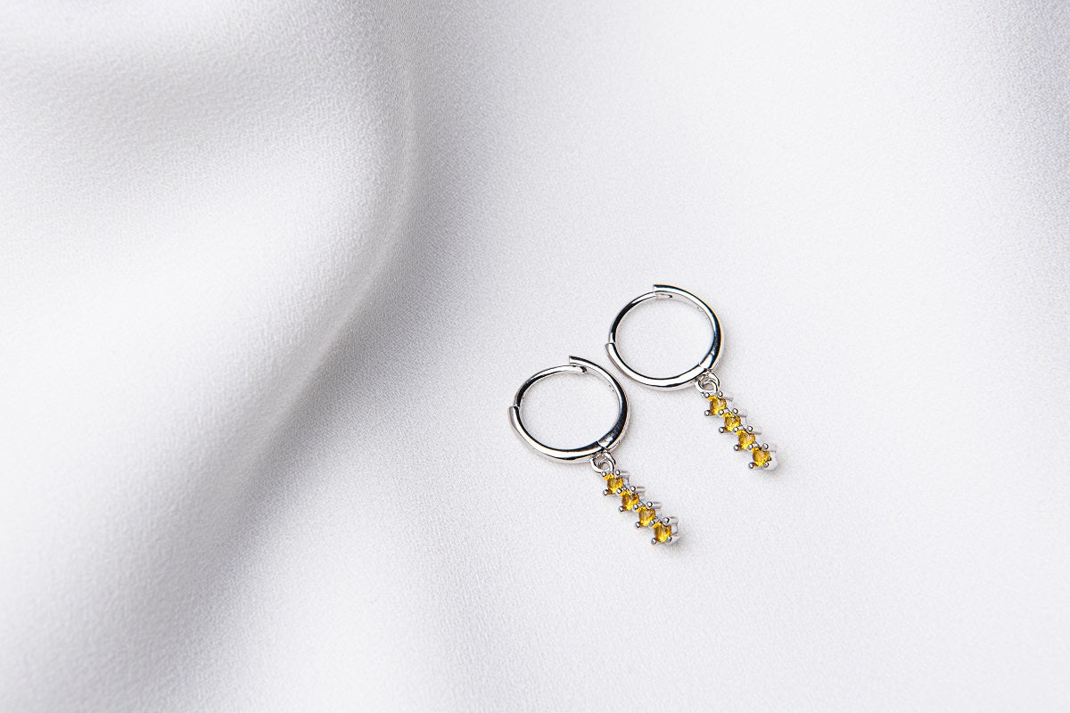 A pair of silver hoop earrings with a dangle of yellow cubic zirconias on a white fabric surface.