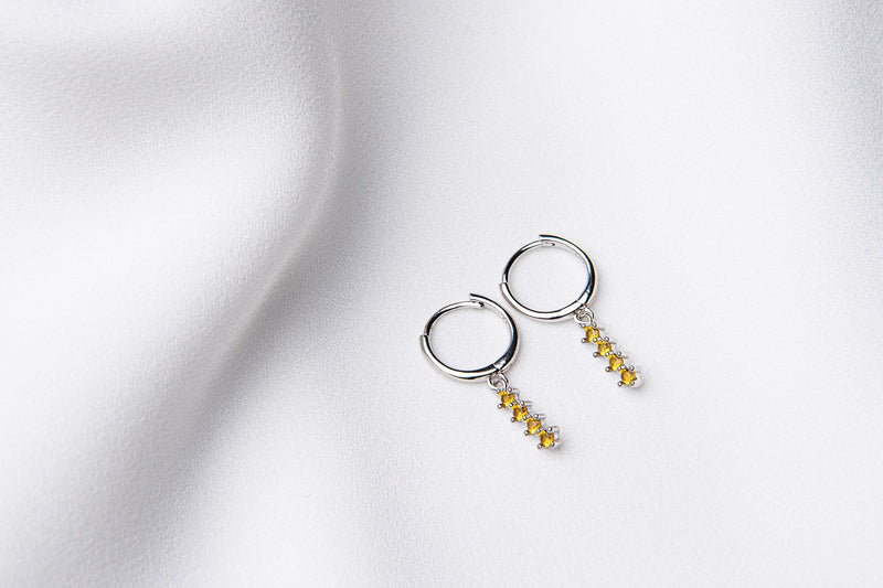 A pair of silver hoop earrings with a dangle of yellow cubic zirconias on a white fabric surface.