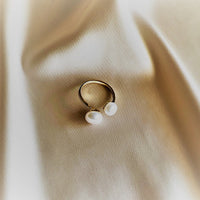 An adjustable gold ring with two white button-shaped pearls placed on a beige fabric surface.