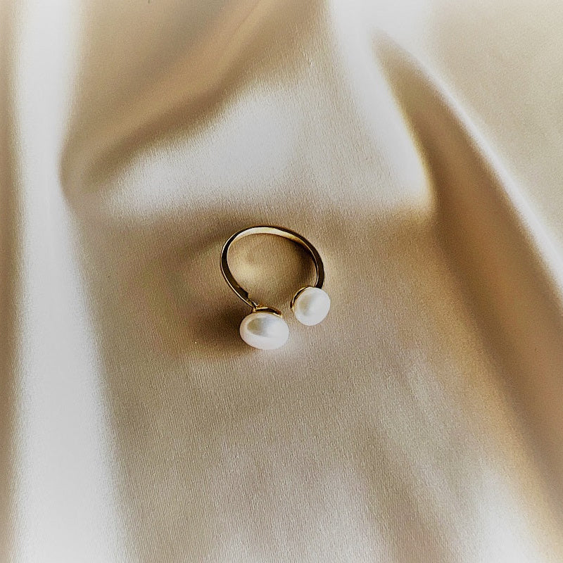 An adjustable gold ring with two white button-shaped pearls placed on a beige fabric surface.