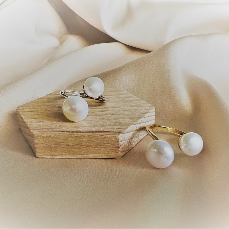 A silver and a gold ring, both featuring two pearls. Set on a wooden and beige background.