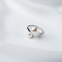 An adjustable silver ring with two white button-shaped pearls placed on a white fabric surface.