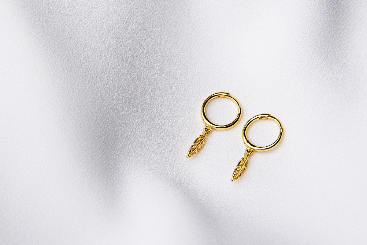 A pair of gold vermeil hoop earrings with feather charms displayed on a white background.