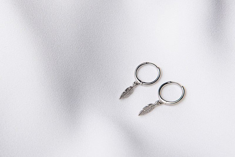 A pair of silver hoop earrings with feather charms displayed on a white background.