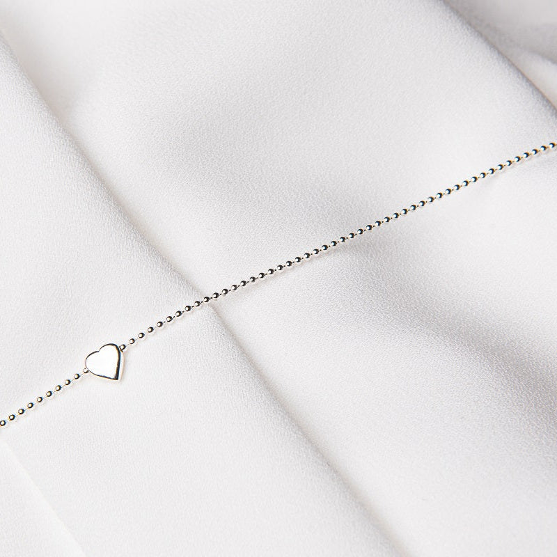 A sterling silver anklet with a beaded chain and a heart-shaped pendant, laid out on a white surface.