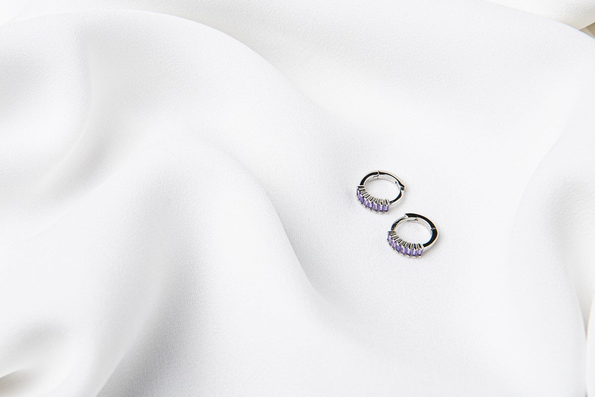 A pair of silver earrings with purple cubic zirconias on a white cloth background