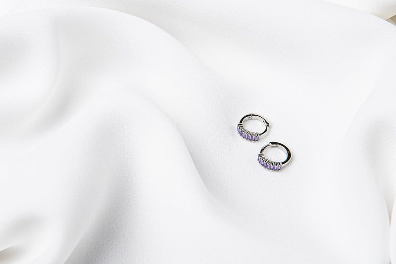 A pair of silver earrings with purple cubic zirconias on a white cloth background
