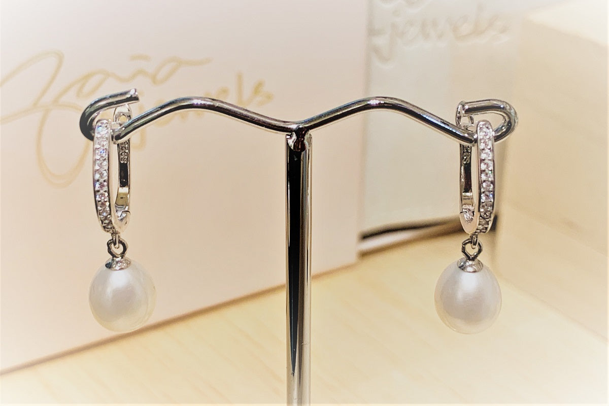 A pair of silver earrlings featureing cubic zirconas and white pearls on a earring display stand with a blurred background