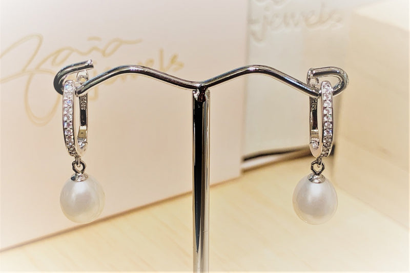 A pair of silver earrlings featureing cubic zirconas and white pearls on a earring display stand with a blurred background