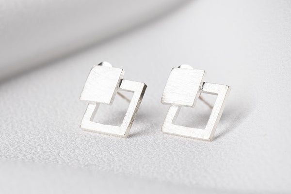 A pair of silver stud earrings with a geometric design, featuring a square within a rectangle, placed on a white surface.