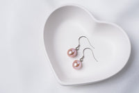 A pair of lavender pearl earrings dangled from silver hooks, placed inside a white, heart-shaped dish.