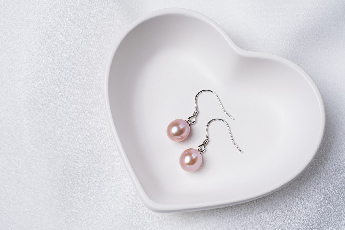 A pair of lavender pearl earrings dangled from silver hooks, placed inside a white, heart-shaped dish.