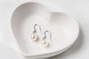 A pair of pearl earrings dangled from silver hooks, placed inside a white, heart-shaped dish.