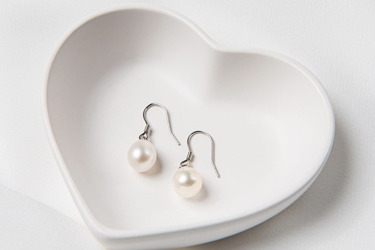 A pair of pearl earrings dangled from silver hooks, placed inside a white, heart-shaped dish.