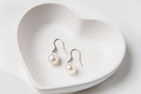 A pair of pearl earrings dangled from silver hooks, placed inside a white, heart-shaped dish.