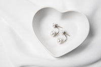 A pair of pearl stud earrings displayed on a white heart-shaped plate, with the back of the earrings showing butterfly backs and a fixed dangle.