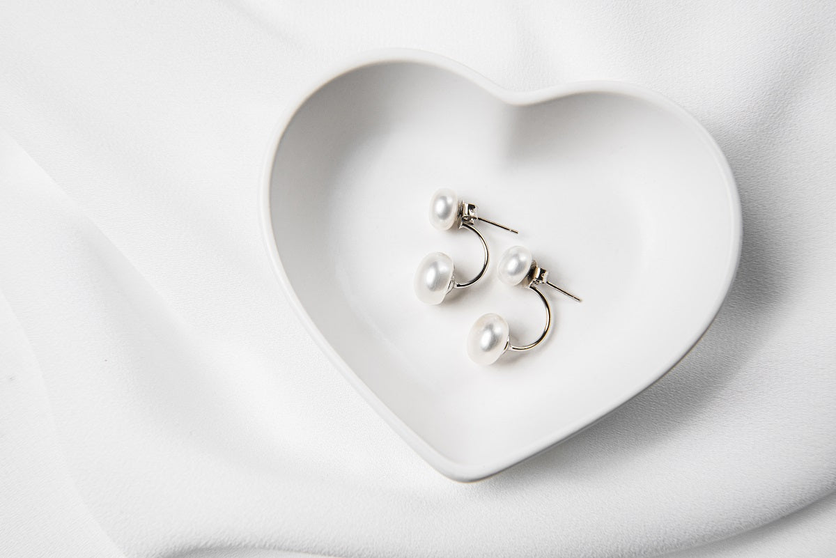 A pair of pearl stud earrings displayed on a white heart-shaped plate, with the back of the earrings showing butterfly backs and a fixed dangle.