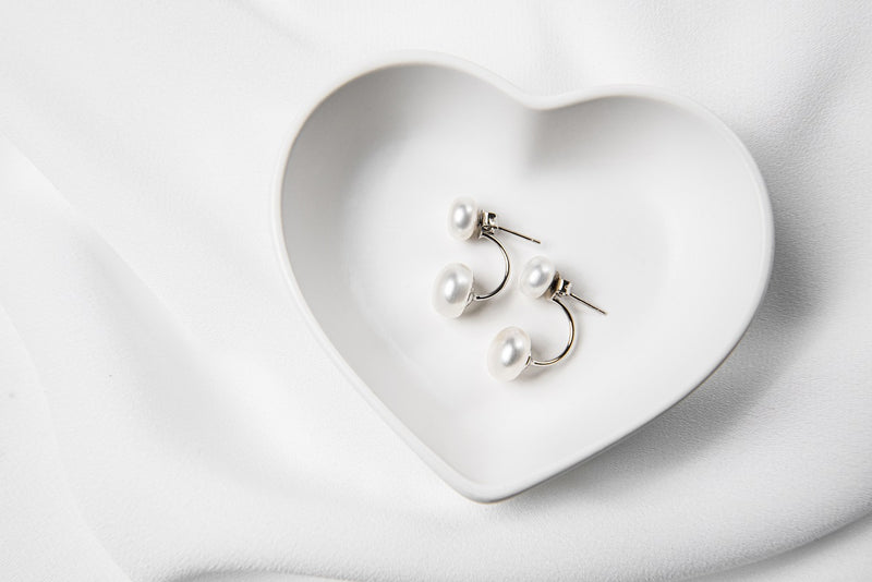 A pair of pearl stud earrings displayed on a white heart-shaped plate, with the back of the earrings showing butterfly backs and a fixed dangle.