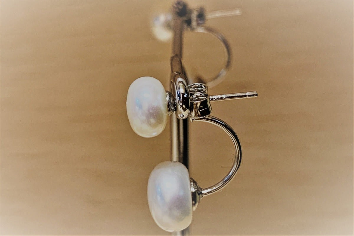 A pair of pearl stud earrings displayed on a stand, with the back of the earrings showing butterfly backs and a fixed dangle.