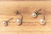 A pair of pearl stud earrings displayed on a wood background, with the back of the earrings showing butterfly backs and a fixed dangle.