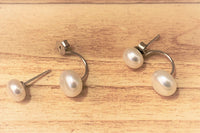 A pair of pearl stud earrings displayed on a wood background, with the back of the earrings showing butterfly backs and a fixed dangle.