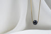 An image of a classic black pearl necklace with an adjustable chain and a single pearl pendant, displayed on a white fabric surface.