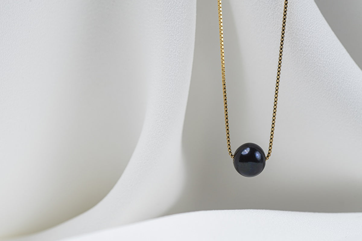 An image of a classic black pearl necklace with an adjustable chain and a single pearl pendant, displayed on a white fabric surface.