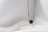 An image of a classic black pearl necklace with an adjustable chain and a single pearl pendant, displayed on a white fabric surface.