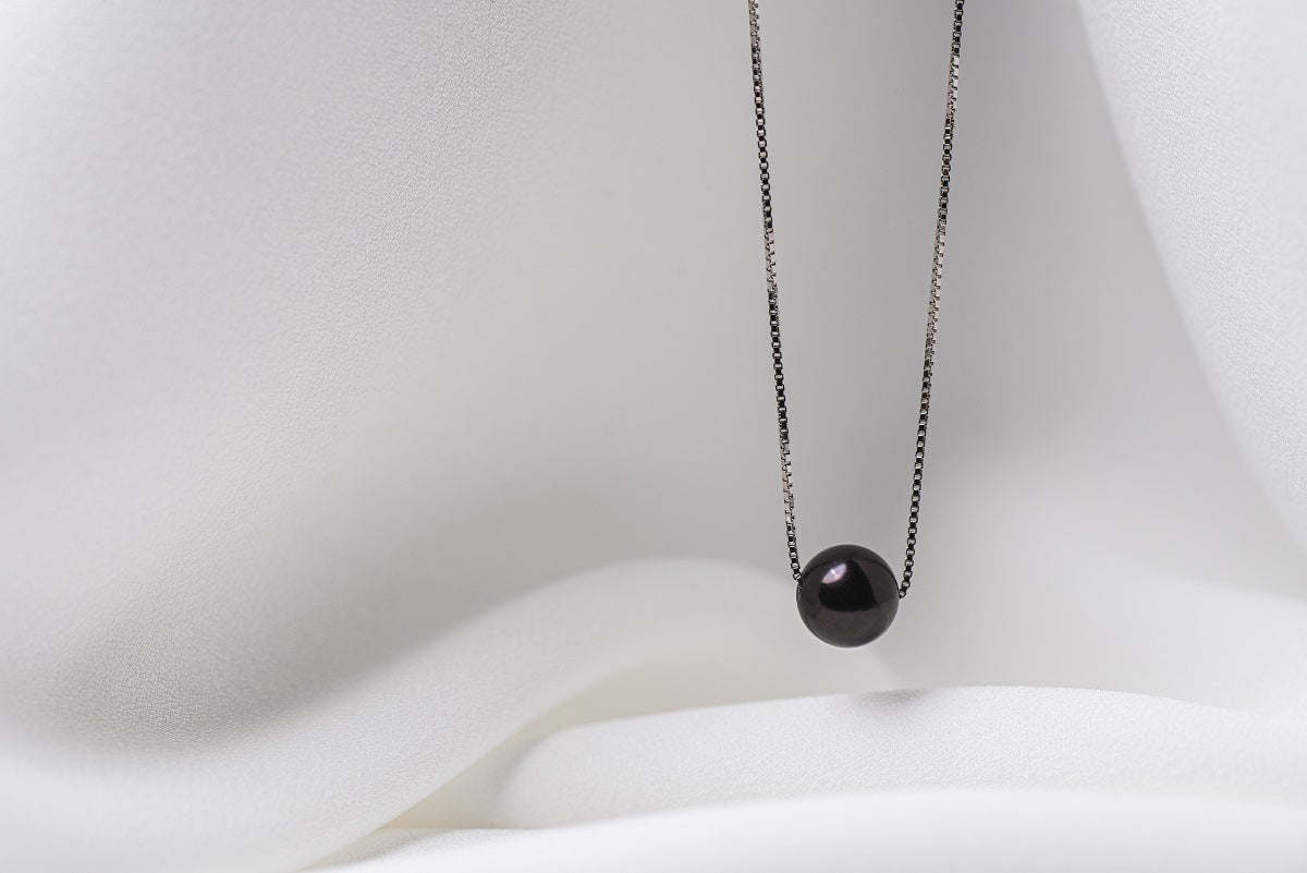 An image of a classic black pearl necklace with an adjustable chain and a single pearl pendant, displayed on a white fabric surface.