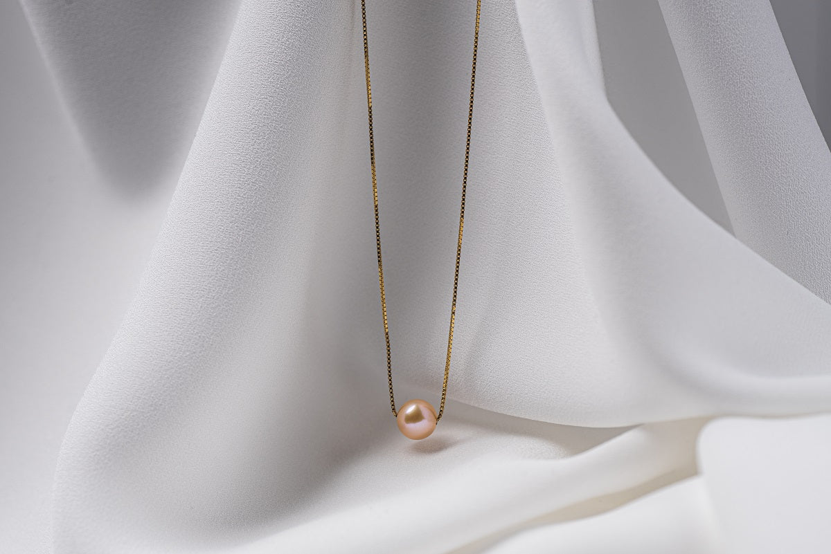 An image of a classic champagne pearl necklace with an adjustable chain and a single pearl pendant, displayed on a white fabric surface.