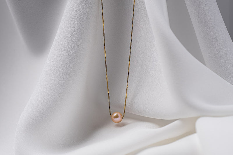 An image of a classic champagne pearl necklace with an adjustable chain and a single pearl pendant, displayed on a white fabric surface.