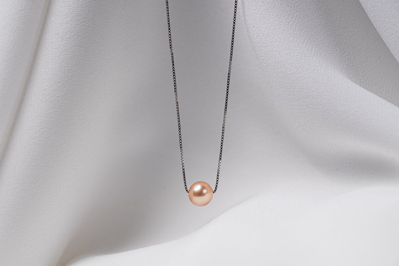 An image of a classic champagne pearl necklace with an adjustable chain and a single pearl pendant, displayed on a white fabric surface.