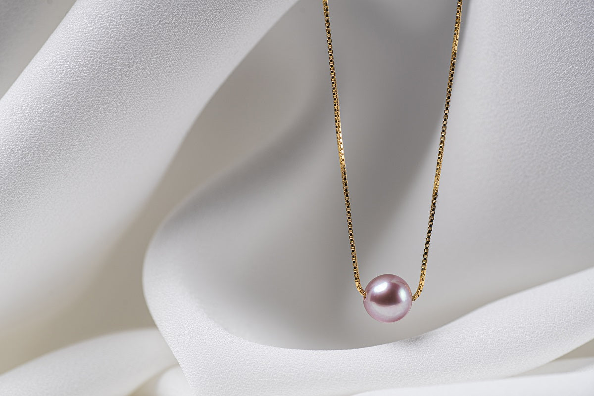 An image of a classic lavender pearl necklace with an adjustable chain and a single pearl pendant, displayed on a white fabric surface.