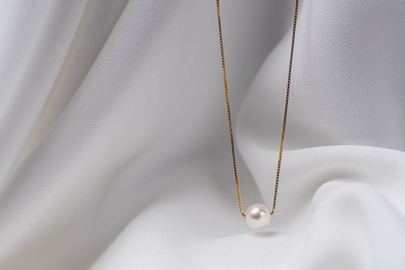 An image of a classic pearl necklace with an adjustable chain and a single pearl pendant, displayed on a white fabric surface.