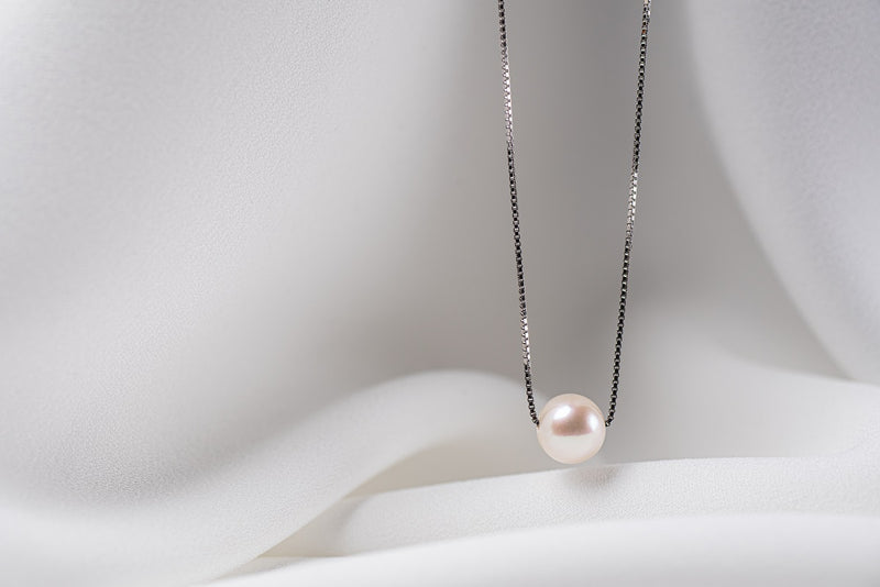 An image of a classic white pearl necklace with an adjustable chain and a single pearl pendant, displayed on a white fabric surface.